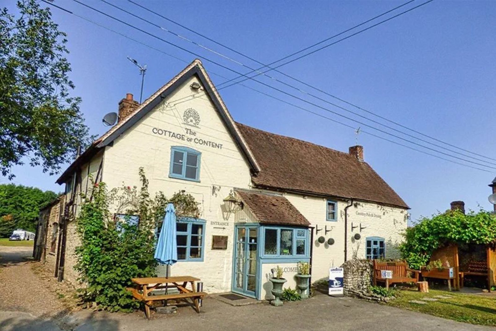 The Cottage of Content - a whitewashed country pub with blue-framed windows, a picnic bench and parasol out front on a sunny day.